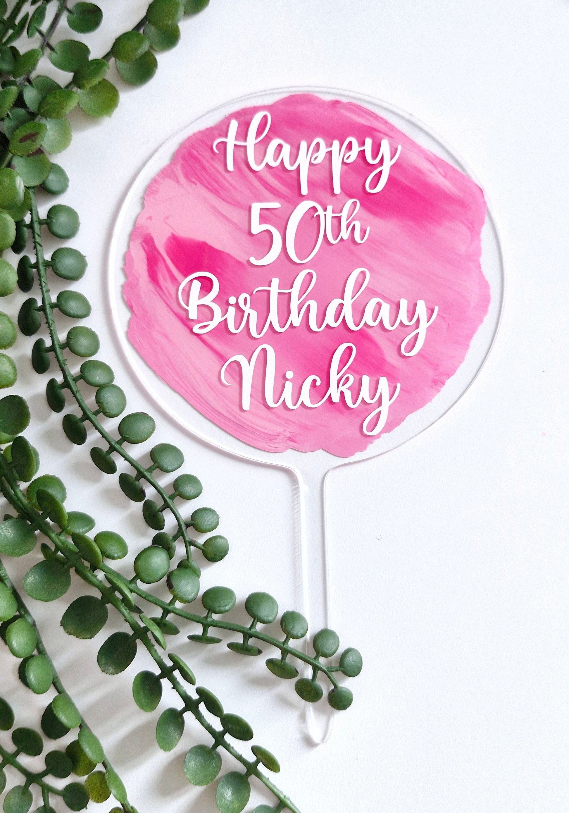 A round birthday cake topper with a pink watercolor design and white text that reads 'Happy 50th Birthday', placed on a cake with greenery decoration.